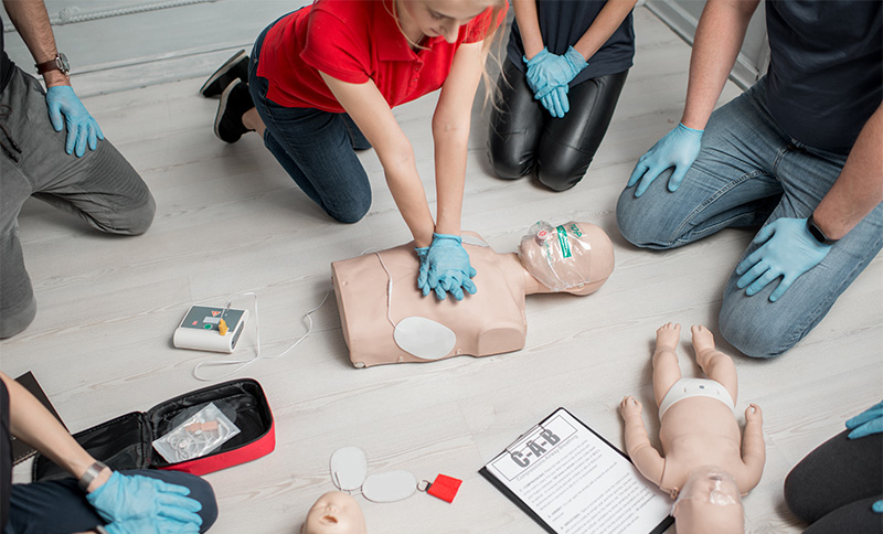 Kvinna visar defibrillering på en HLR docka för en grupp som sitter i en rund ring.
