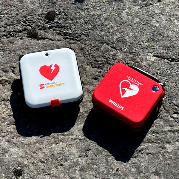 Två defibrillatorer, en vit LIFEPAK CR2 och en röd Philips Heartstart, ligger på en grov stenyta.