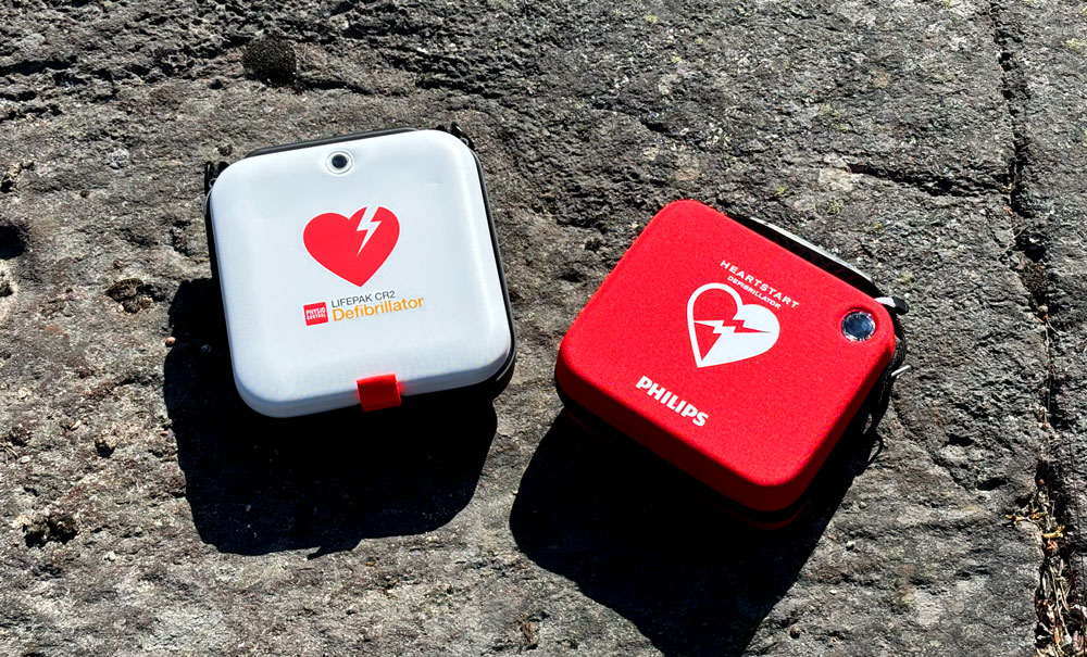 En vit LIFEPAK CR2 hjärtstartare och en röd Philips Heartstart hjärtstartare ligger sida vid sida på en grå, ojämn stenyta.