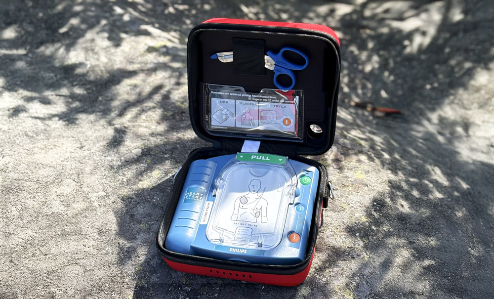 En Philips Heartstart HS1-hjärtstartare ligger öppen i sin röda väska på en stenig yta. Väskan innehåller sax, rakblad och instruktioner för användning.