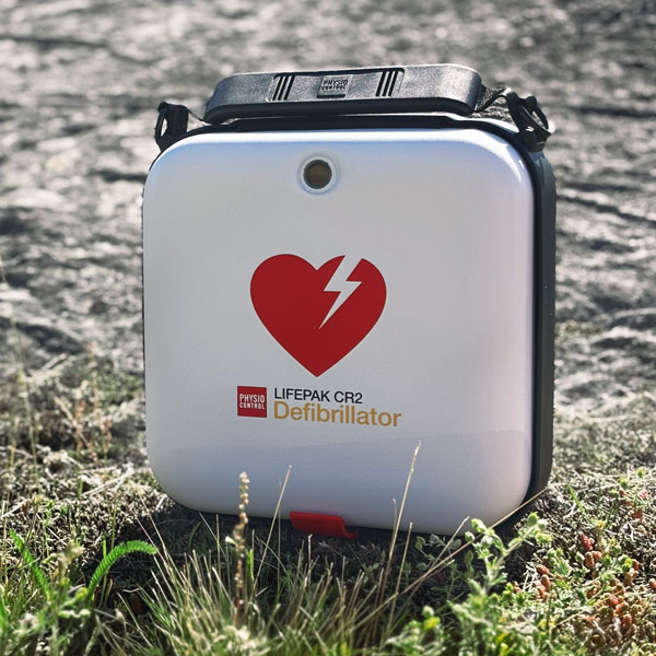 En vit LIFEPAK CR2 defibrillator med ett rött hjärtlogo står upprätt på en stenig mark med grön vegetation.