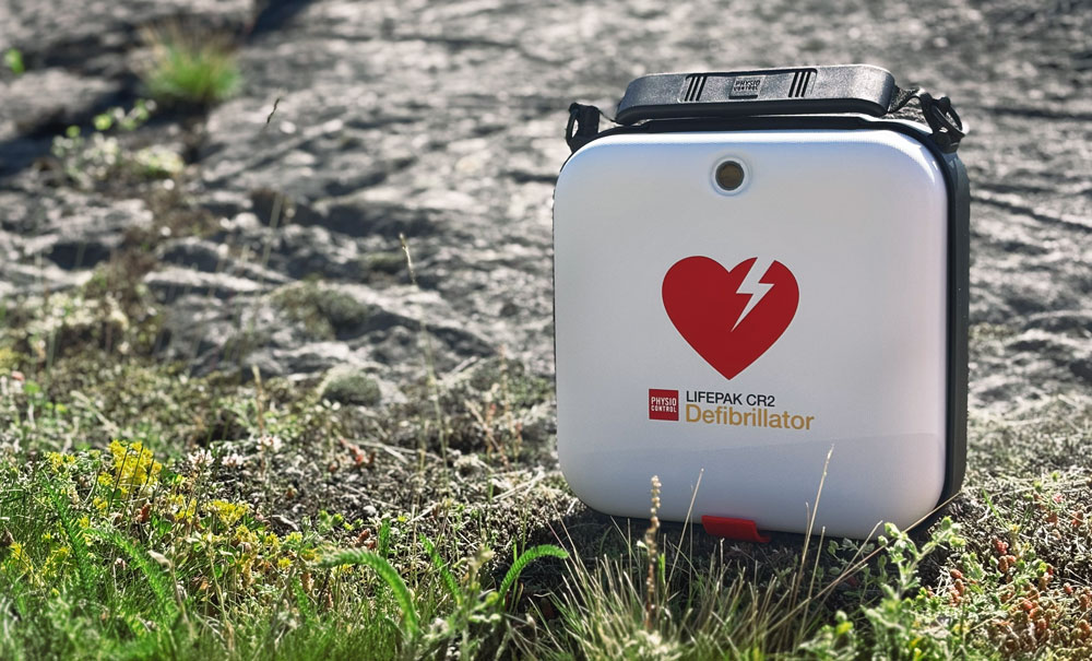 En vit LIFEPAK CR2 hjärtstartare med ett rött hjärtlogo står i gräset framför en bergig yta.