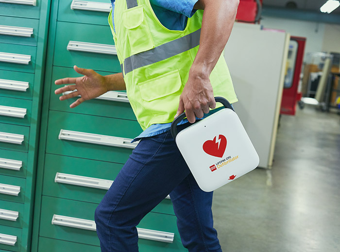 Kroppen av en man springandes med gul reflexväst hållandes i hjärtstartaren Lifepak CR2 i ena handen inne i en lagerlokal. 