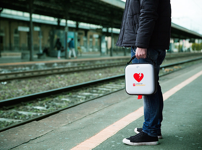 En man som håller i en hjärtstartare av modell Lifepak CR2. Mannen står ute på en tågperrong.