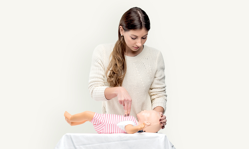 Hjärtkompressioner med två fingrar på en dockbebis i en vit-rosa sparkdräkt som utförs av en brunhårig kvinna i en vit stickad tröja. 
