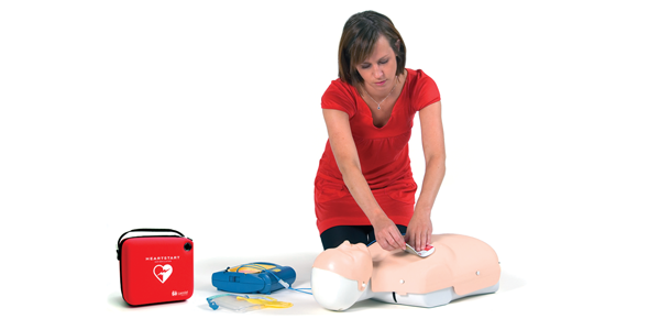 Kvinna som sätter på elektroder på en halvkropp HLR-docka. På golvet bredvid ligger en övningshjärtstartare med tillhörande väska. 